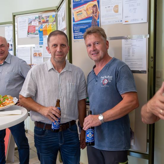 Drei Männer stehen vor einer Pinnwand mit verschiedenen angehefteten Papieren. Der Mann in der Mitte hält eine Bierflasche. Der Mann auf der rechten Seite trägt ein blaues Shirt mit Logo.