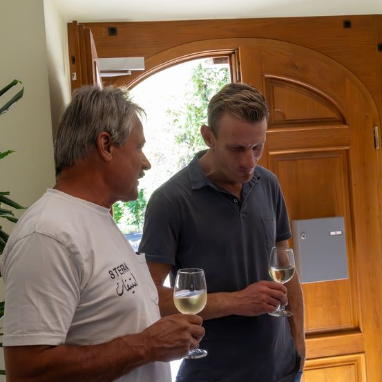 Zwei Männer, einer mit grauem Hemd und der andere in Weiß, probieren Wein vor einer Holztür.