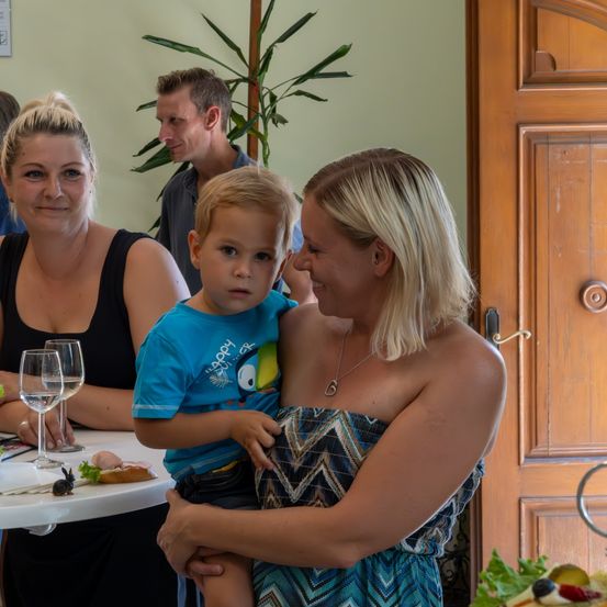 Eine Familie sitzt an einem Tisch mit einem kleinen Jungen, der die Hand seiner Mutter hält. Zwei Frauen und zwei Männer lächeln. Im Hintergrund befindet sich eine Holztür und eine Pflanze.