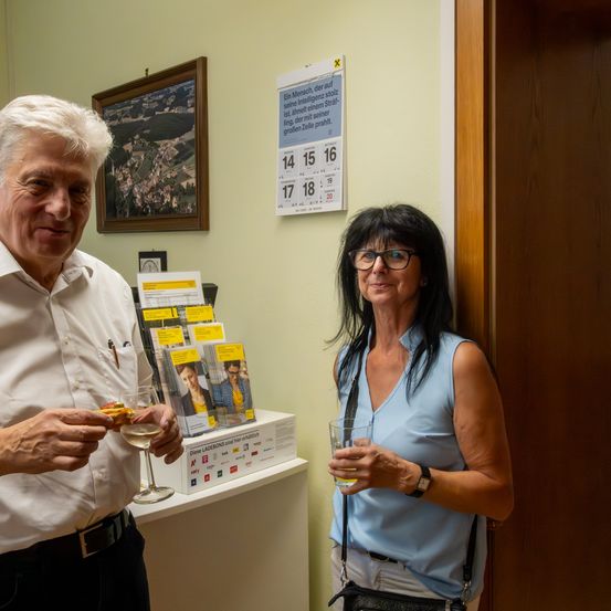 Ein älterer Mann und eine Frau stehen in einem Raum und lächeln. Der Mann hält ein Glas Wein, während die Frau ein Glas mit einem Getränk in der Hand hat. Hinter ihnen ist ein Kalender an der Wand befestigt, und ein gerahmtes Bild hängt über einem Regal mit Broschüren.