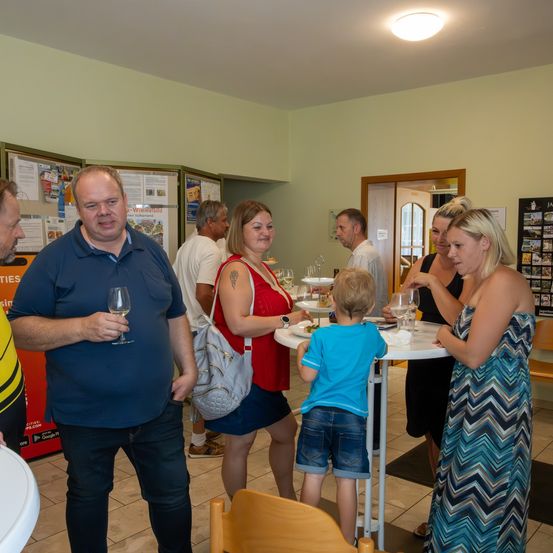 Eine Gruppe von Erwachsenen und Kindern versammelt sich um einen Tisch mit Weingläsern und Tellern, vielleicht für eine gesellige Veranstaltung. Ein Kind steht und hält ein Weinglas. Der Raum hat einen gefliesten Boden und Wände mit gerahmten Bildern und Postern.