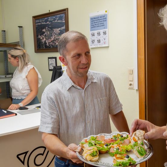 Ein Mann in einem gestreiften Hemd hält einen Tablett mit Sandwiches, während eine Frau hinter ihm an einem Schreibtisch arbeitet. Ein Kalender und ein gerahmtes Bild hängen an der Wand.