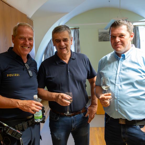 Drei Männer stehen lächelnd in einem Raum. Der Mann auf der linken Seite trägt eine Polizeiuniform, während der Mann in der Mitte ein Weinglas hält. Der Mann auf der rechten Seite trägt ein blaues Hemd und hält ebenfalls ein Weinglas.