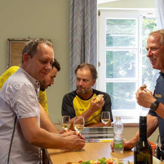 Vier Männer sitzen an einem Tisch mit Essen und Weingläsern, lächeln und lachen. Der Mann im gelben Shirt hält ein Sandwich, während der Mann im grauen Shirt ein Stück Brot hält. Der Tisch hat Teller, Weingläser und eine Flasche Wein. Hinter ihnen ist ein Fenster mit einem Vorhang.