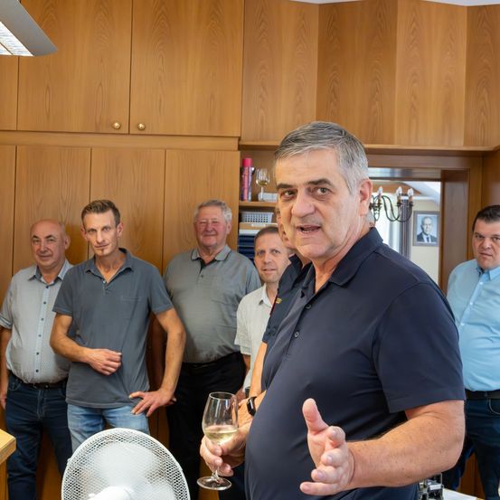 Eine Gruppe von Männern steht in einem Raum, wobei einer vorn einen Daumen nach oben gibt und ein Glas Wein in der Hand hält. Sie sind alle in Freizeitkleidung gekleidet.