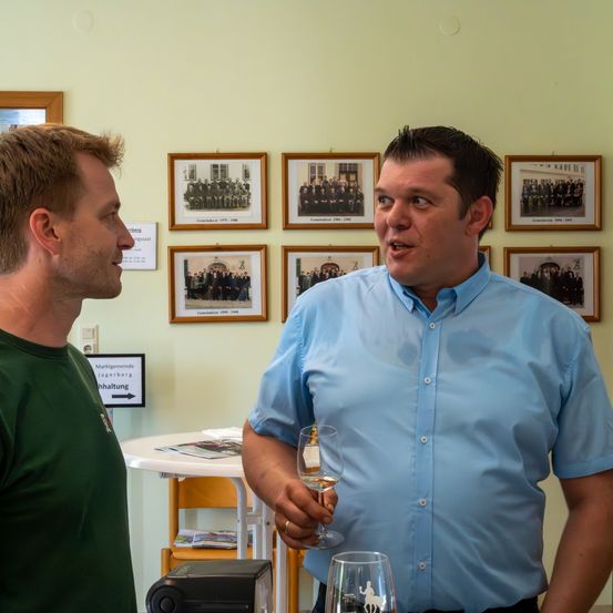 Zwei Männer stehen vor einer Wand mit gerahmten Bildern, einer hält ein Weinglas.