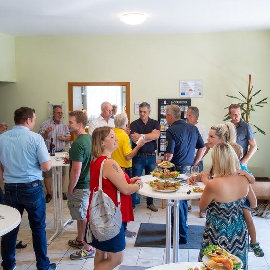 Eine Gruppe Erwachsener in Freizeitkleidung steht um Tische herum, unterhält sich und isst. Sie befinden sich in einem Raum mit gelben Wänden und einer Pflanze in der Ecke. Eine Frau in einem roten Shirt hält einen Tablett mit Essen.