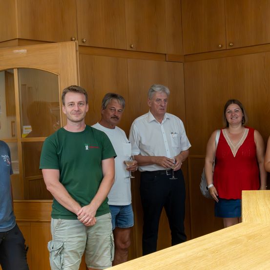 Eine Gruppe von fünf Personen steht in einem Raum. Der Mann in Grün lächelt, während die anderen ernst aussehen. Sie alle halten Weingläser. Die Frau in Rot trägt eine Halskette. Hinter ihnen befinden sich Holzschränke.