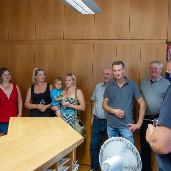 Eine Gruppe von Menschen steht in einem Raum mit Holzschränken. Einige halten Weingläser. Ein Ventilator und ein Holztisch sind im Vordergrund.