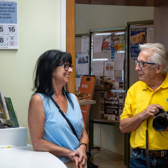 Eine Frau in einem blauen ärmellosen Oberteil und ein Mann in einem gelben Hemd lächeln und unterhalten sich. Dahinter befindet sich ein Aushang mit verschiedenen Papieren und einem Kalender an der Wand.