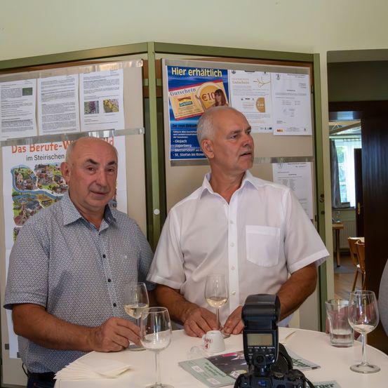 Zwei Männer sitzen an einem Tisch mit Weinläsern und lächeln für ein Foto. Hinter ihnen wirbt ein Plakat für einen 10-Euro-Gutschein.