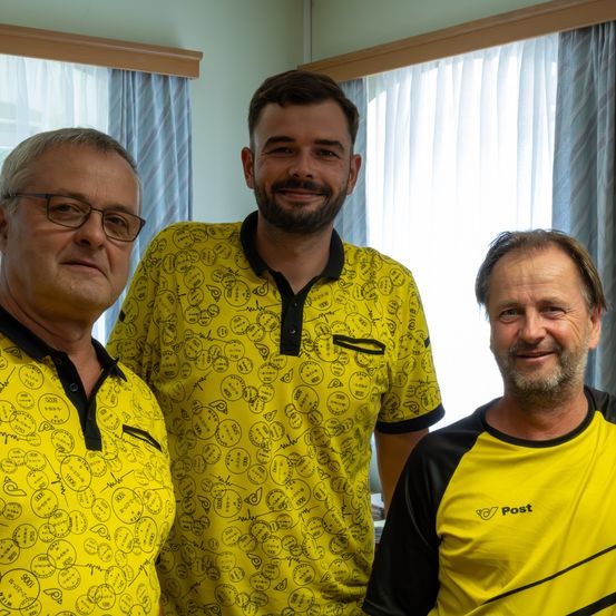Drei Männer in gelben Polo-Shirts mit verschiedenen Designs, eines mit einem Logo auf der linken Brust, stehen zusammen vor einem Fenster mit blauen Vorhängen.