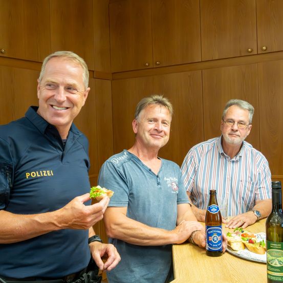Drei Männer sitzen lächelnd am Tisch. Einer hält ein Sandwich, ein anderer hat eine Bierflasche und der dritte trägt eine Polizeiuniform. Hinter ihnen befinden sich Holzschränke.