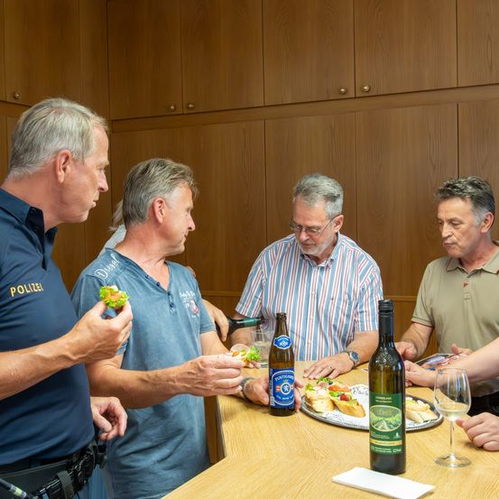 Fünf Männer versammeln sich um einen Tisch mit Essen und Wein. Ein Mann in einer blauen Uniform mit dem Wort 'POLIZEI' darauf hält ein Sandwich. Ein anderer Mann hält eine Flasche Wein.