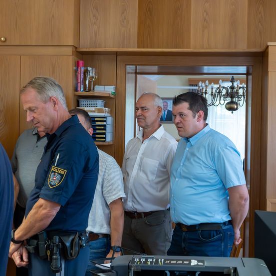 Eine Gruppe von Männern, möglicherweise Polizeibeamte, steht in einem Raum. Ein Beamter hat ein Abzeichen auf der Schulter. Vor ihnen steht ein Mann in einem blauen Hemd und ein Mann in einem weißen Hemd. Hinter ihnen befindet sich ein Spiegel und eine Deckenleuchte.