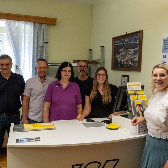 Eine Gruppe von Menschen steht lächelnd hinter einem Schreibtisch und posiert für ein Foto. Eine Frau trägt ein lila Shirt, eine andere schwarz und eine dritte weiß. Ein Kalender hängt an der Wand.