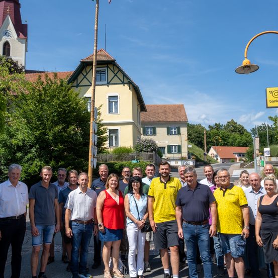 Eine Gruppe von Menschen steht vor einem gelben Gebäude unter blauem Himmel. Sie sind lässig gekleidet und scheinen für ein Foto zu posieren. Ein Fahnenmast steht prominent im Vordergrund.