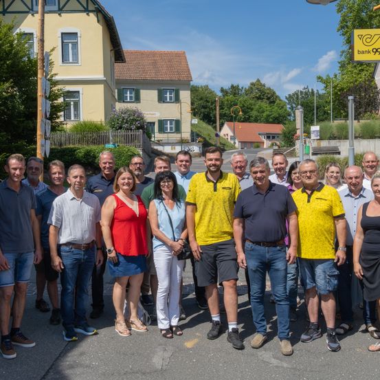 Eine Gruppe von Menschen posiert für ein Foto auf einer Straße. Sie stehen in einer Reihe, lächeln und tragen Freizeitkleidung. Der Hintergrund zeigt Häuser und eine Straße.