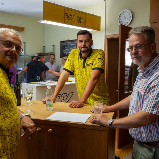 Drei Männer stehen in einem Raum. Ein Mann trägt ein gelbes Shirt mit Logo. Ein anderer trägt ein gestreiftes Shirt. Vor ihnen steht ein Tisch mit Weingläsern und einer Flasche. Hinter ihnen stehen und sitzen Menschen. Eine Uhr und ein Bilderrahmen hängen an der Wand.