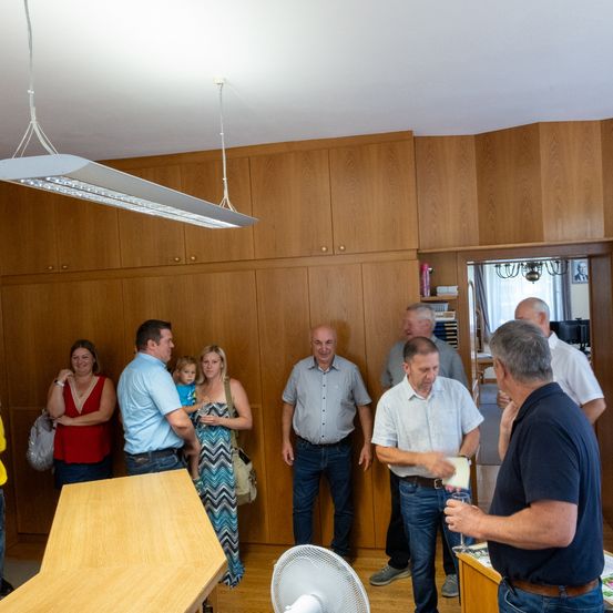 Eine Gruppe von Menschen steht in einem Raum mit Holzschränken und einer Laterne. Ein Mann hält ein Stück Papier und ein Glas. Ein anderer Mann in einem blauen Shirt trägt ein Kind.