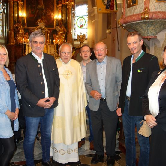 Sechs Menschen stehen lächelnd für ein Foto in einer Kirche. Der Mann in der Mitte trägt eine weiße Robe. Die anderen sind in Anzügen und Freizeitkleidung gekleidet. Hinter ihnen steht ein Altar mit Kerzen.