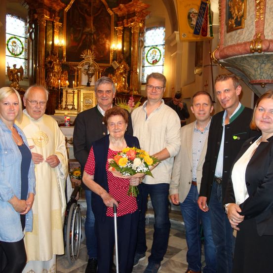 Eine Gruppe von Menschen steht lächelnd in einer Kirche. Eine ältere Frau hält einen Blumenstrauß, während ein Priester hinter ihr steht. Zwei Männer in Anzügen flankieren sie, und andere stehen in der Nähe. Die Kirche hat Buntglasfenster und einen großen Altar.