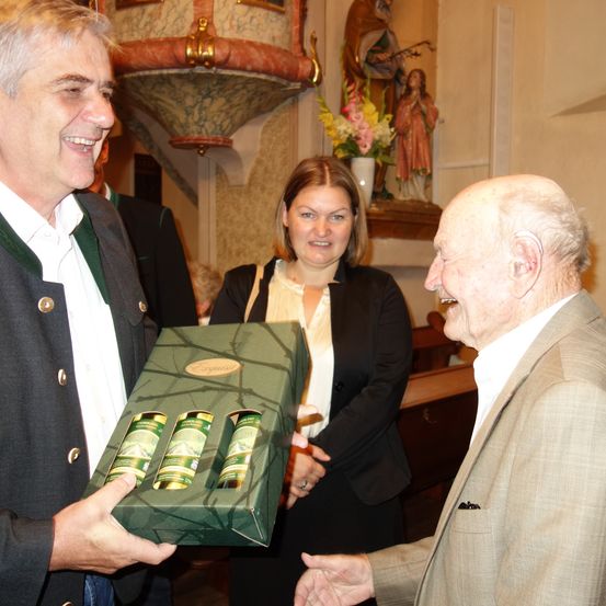 Ein Mann überreicht einem älteren Mann in einer Kirche eine Geschenkbox. Eine Frau steht hinter ihnen.