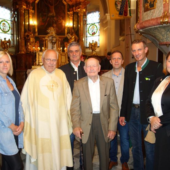 Eine Gruppe von Menschen steht in einer Kirche. Einer von ihnen ist ein Priester in einem goldenen Gewand. Ein älterer Mann hält einen Stock.