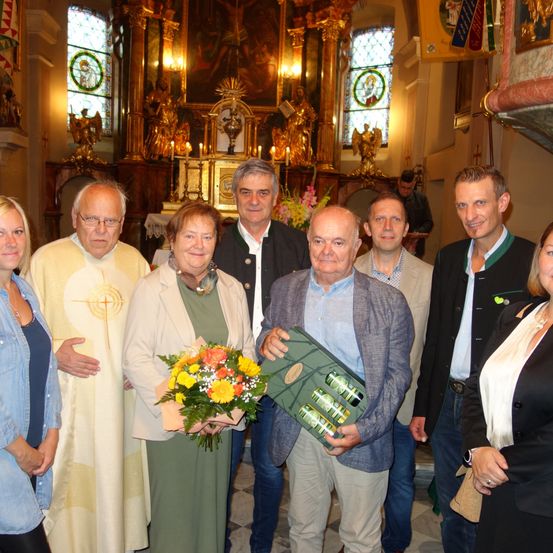 Eine Gruppe von Menschen steht in einer Kirche und lächelt für ein Foto. Die Frau in der Mitte hält einen Blumenstrauß und eine Geschenkbox. Hinter ihnen steht ein Altar mit einer Statue und Kerzen.