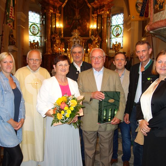 Eine Gruppe von Menschen steht lächelnd in einer Kirche und posiert für ein Foto. Eine ältere Frau hält einen Blumenstrauß und ein Mann einen grünen Kasten. Dahinter steht ein Priester.