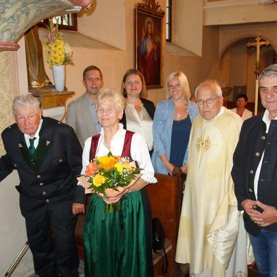 Eine Gruppe von Menschen steht in einer Kirche und lächelt für ein Foto. Eine ältere Frau hält einen Blumenstrauß. Ein Priester ist anwesend und ein religiöses Gemälde schmückt die Wand.