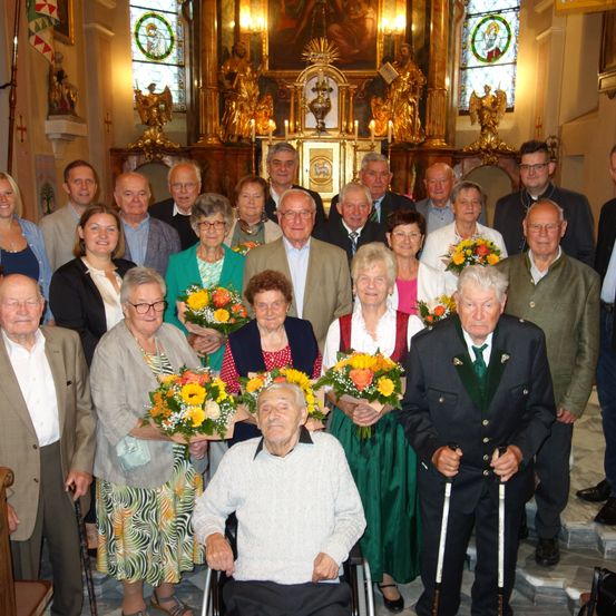 Eine Gruppe älterer Menschen versammelt sich in einer Kirche, einige halten Blumen. Ein älterer Mann im Rollstuhl steht in der Mitte. Hinter ihnen befinden sich goldene Statuen, eine Statue von Jesus und ein Kreuz.