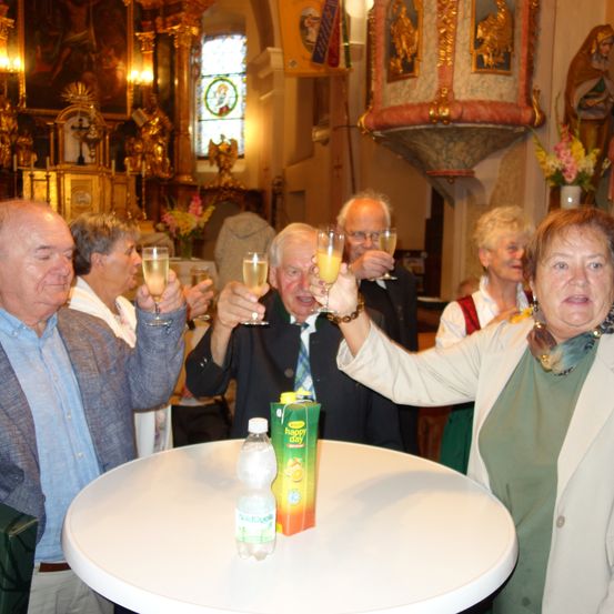 Eine Gruppe älterer Erwachsener stoßen mit Weinläsern in einer Kirche an. Ein Tisch mit einer Flasche und einer Schachtel Saft im Vordergrund.