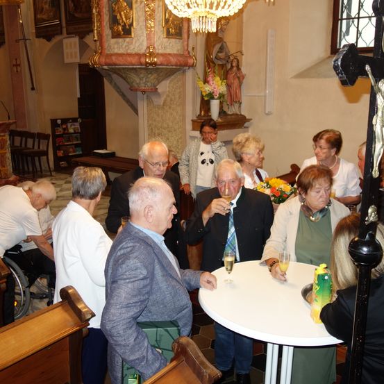 In einer Kirche versammeln sich ältere Menschen, einige stehen, andere sitzen. Ein Mann in Anzug trinkt aus einem Weinglas. Die Kirche ist mit religiösen Ikonen und einer Kronleuchter dekoriert.