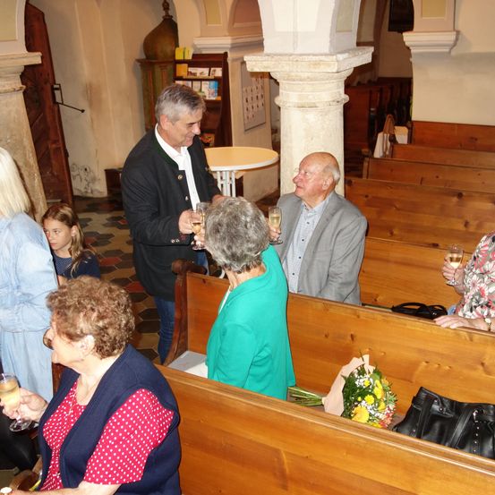 Mehrere Menschen versammeln sich in einer Kirche und halten Weingläser. Ein älteres Ehepaar stößt an, während andere sitzen und stehen. Im Hintergrund sind ein Tisch und eine Bank zu sehen.