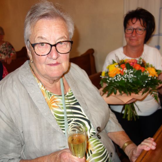 Eine ältere Frau hält ein Glas Champagner, während eine andere Frau einen Blumenstrauß im Hintergrund hält.