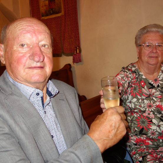 Ein älterer Mann und eine ältere Frau sitzen und lächeln. Der Mann hält ein Glas Champagner, während die Frau eine Brille trägt und ein Blumenkleid anhat. Hinter ihnen zieren ein roter Vorhang und ein Bilderrahmen die Wand.