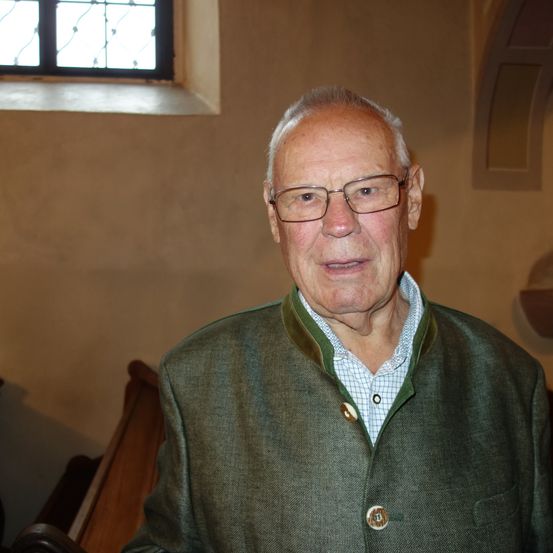 Ein älterer Mann mit Brille und grüner Jacke steht in einer Kirche.