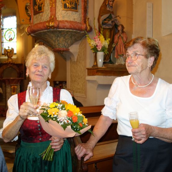 Zwei ältere Frauen stehen in einer Kirche, eine hält einen Blumenstrauß und Champagner, die andere ein Glas Orangensaft. Hinter ihnen steht eine Statue einer Heiligen.