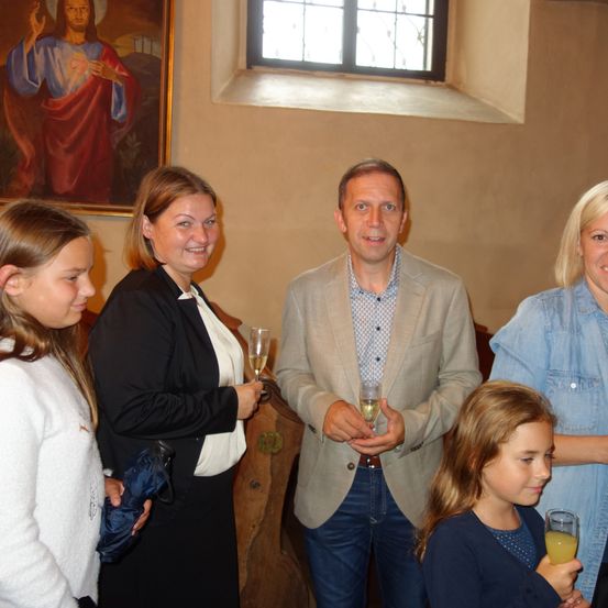 Eine Gruppe von Menschen in einem Raum, lächelnd und Champagnerflöten haltend. Eine Frau hält einen blauen Regenschirm. Hinter ihnen eine Wand mit einem Gemälde und einem Fenster.