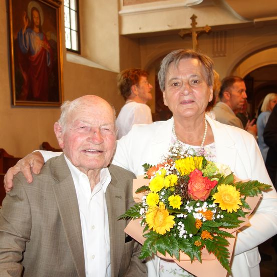 Ein älteres Paar steht in einer Kirche. Der Mann trägt einen Anzug, und die Frau hält einen Blumenstrauß. Hinter ihnen sind ein religiöses Gemälde und ein Kreuz zu sehen, mit Menschen im Hintergrund.