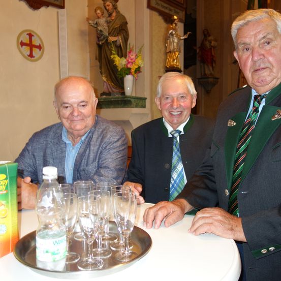 Drei ältere Männer sitzen um einen Tisch in einer Kirche. Sie lächeln und scheinen für ein Foto zu posieren. Es gibt Weingläser, eine Flasche und eine Schachtel mit etwas auf dem Tisch. Hinter ihnen befinden sich Statuen von Heiligen und ein Kreuz an der Wand.