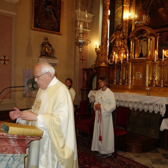 Ein Priester steht am Altar und spricht in ein Mikrofon. Hinter ihm hält eine Frau eine rote Schnur. Im Hintergrund steht eine Statue eines Heiligen und Kerzen auf einem goldenen Altar.