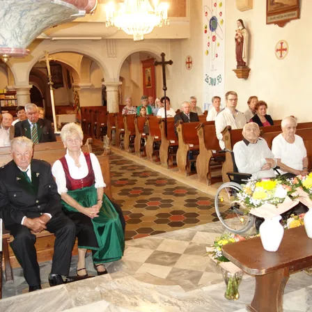 Eine Indoor-Kirchenversammlung mit älteren Teilnehmern. Ein Mann und eine Frau sitzen vorne. Kirchenbänke säumen die Seiten, und ein Rollstuhl steht in der Nähe von Blumen.