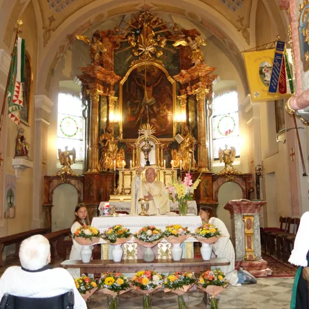 Ein Priester steht hinter einem Altar in einer Kirche mit vielen Blumen, einer Flagge und einem Gemälde. Zwei Frauen knien vor dem Altar, und zwei Personen sitzen auf den Kirchenbänken.