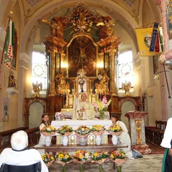 Ein Priester steht hinter einem Altar in einer Kirche mit vielen Blumen, einer Flagge und einem Gemälde. Zwei Frauen knien vor dem Altar, und zwei Personen sitzen auf den Kirchenbänken.