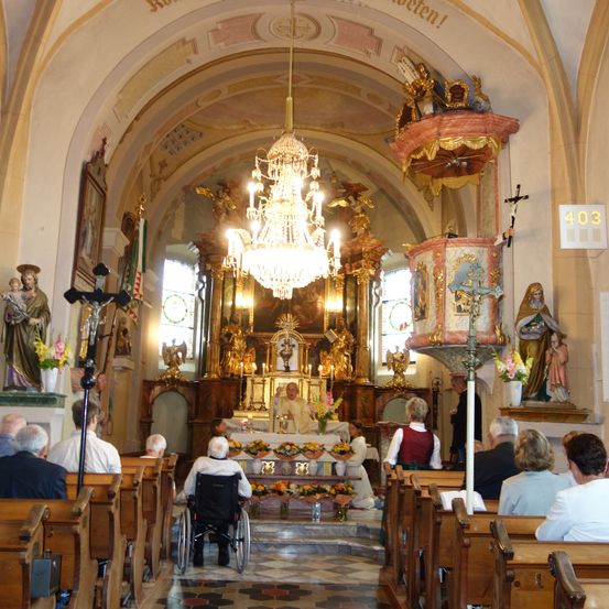 Das Innere einer Kirche zeigt einen Altar mit einem Priester, Kerzen und Blumen. Die Bänke sind mit Gottesdienstbesuchern gefüllt, und Statuen schmücken die Wände. Ein Kronleuchter hängt von der Decke.