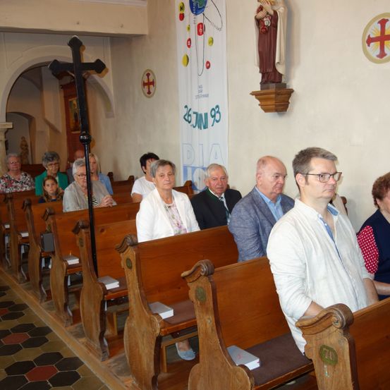 Eine Gruppe von Menschen in einer Kirche, die einen Gottesdienst besuchen, einige sitzen auf Bänken, andere stehen. Der Innenraum ist mit Holzbänken, einem Kreuz und religiösen Dekorationen ausgestattet.