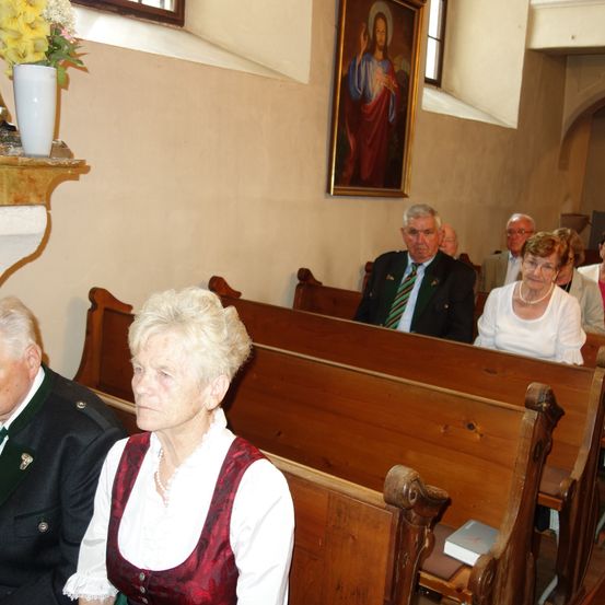 Eine Gruppe älterer Menschen sitzt in einer Kirche. Ein Mann und eine Frau in traditioneller Kleidung sitzen in der vorderen Reihe.