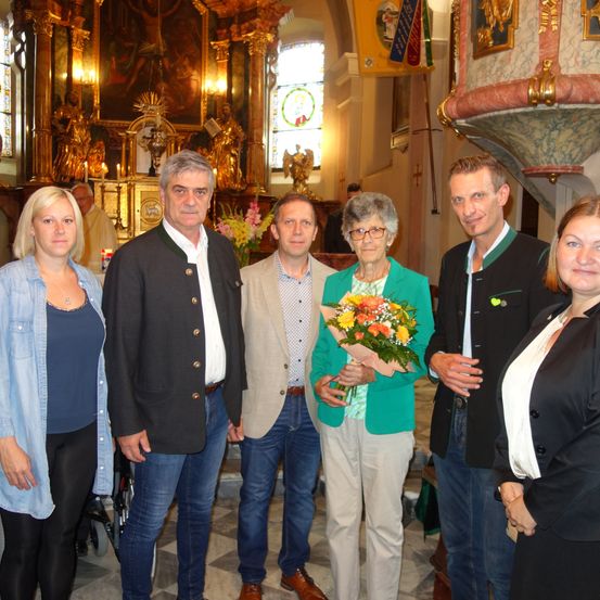 Eine Gruppe von Menschen, darunter eine Frau mit Brille, die einen Blumenstrauß hält, steht in einer Kirche mit kunstvollen Dekorationen und Buntglasfenstern.